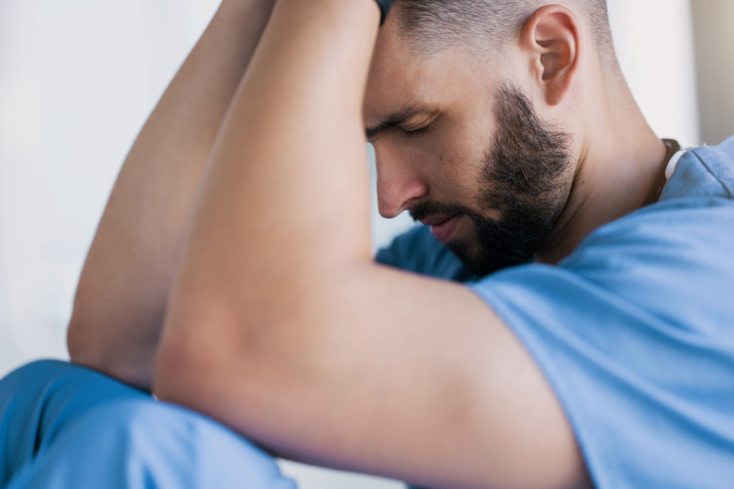 Treating fibromyalgia, image of a man wearing a blue shirt and blue pants, sitting with his eyes closed, head tilted forward with his hands placed on top of his head.