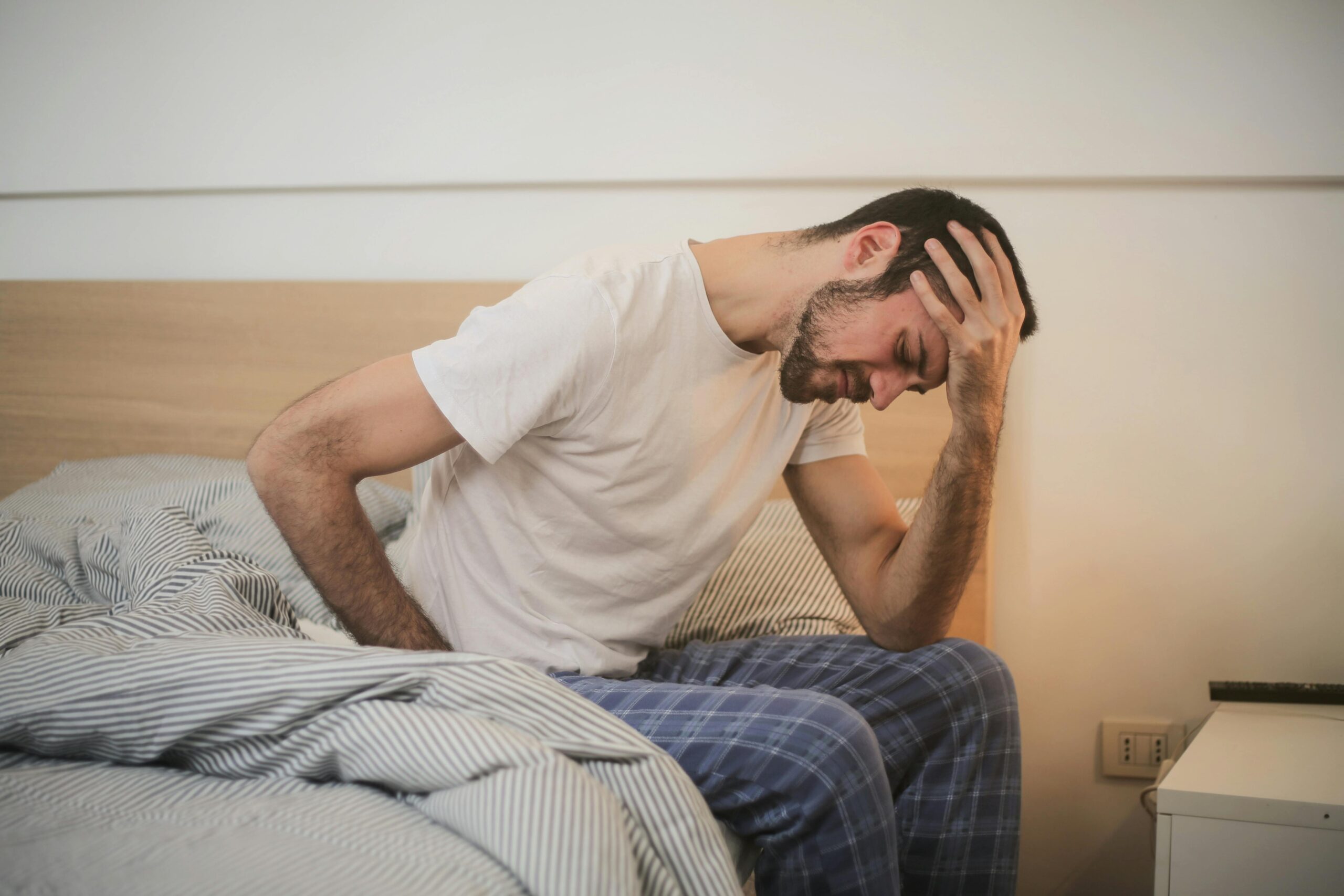 Treating chronic fatigue syndrome, image of a man sitting on the edge of his bed, holding his head with his left hand and a look of pain on his face.