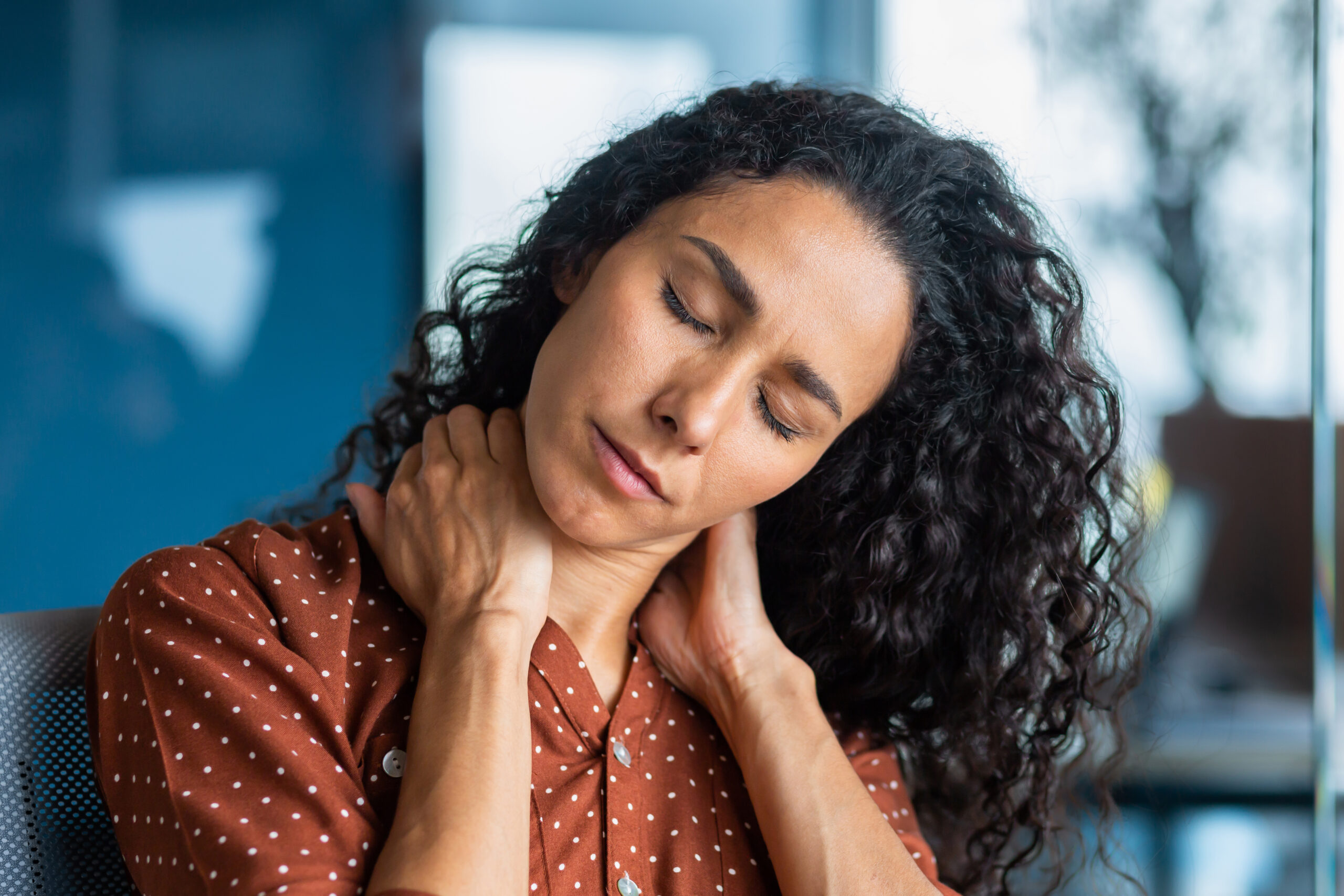 Treating fibromyalgia, image of a woman with long, dark, curly hair and her eyes closed, grasping her neck with both of her hands.