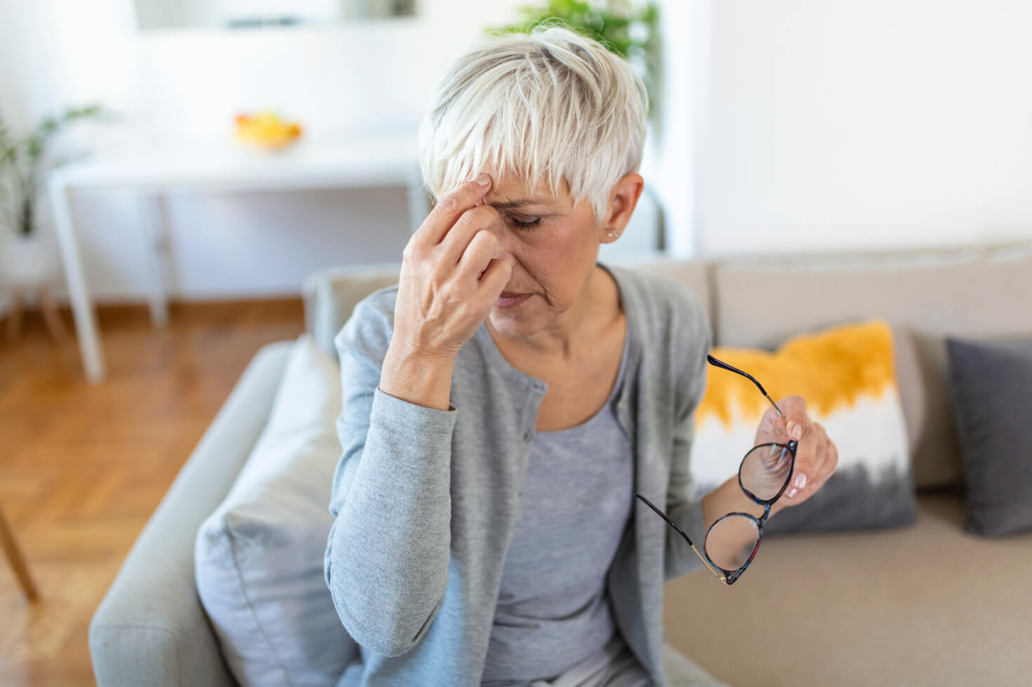 Headaches and Chronic Migraines, image of a woman sitting on the edge of a gray couch holding her glasses in her left hand and touching her forehead with her right hand.