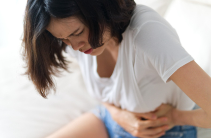 Polycystic Ovary Syndrome (PCOS), image of a woman sitting on a white bed, grasping her stomach with both hands with a look of pain across her face.