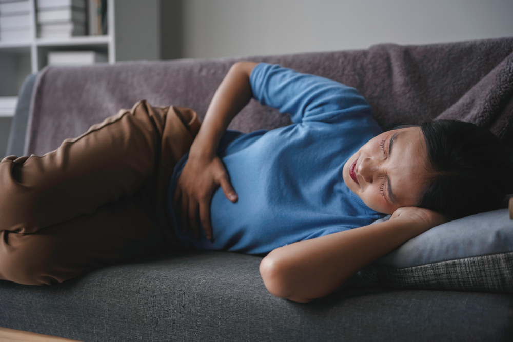 Inflammatory Bowel Disease (IBD), image of a young woman wearing a blue long sleeve shirt and brown pants lying on the couch in pain and grabbing her stomach with her right hand.