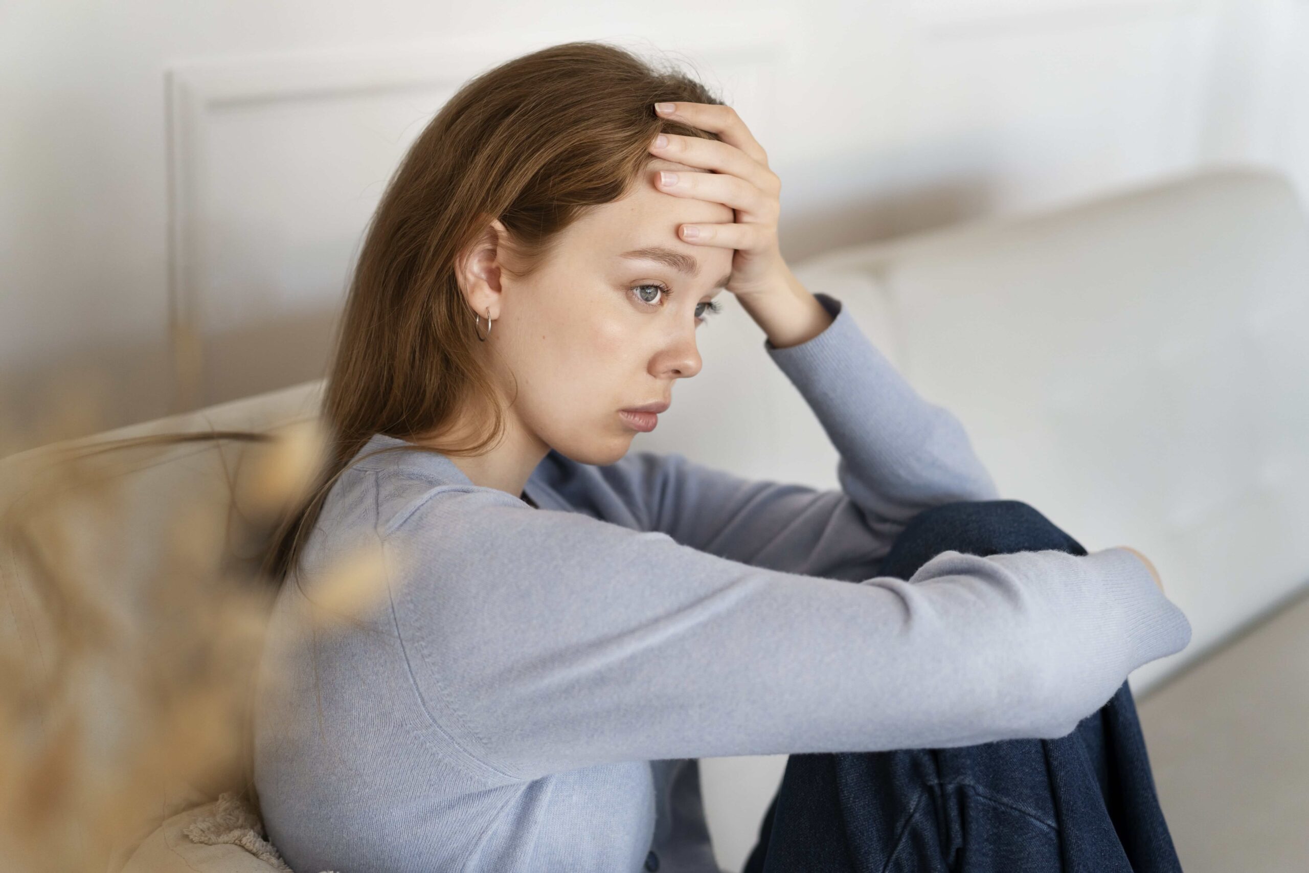Chronic Anxiety, image of a sad woman sitting on a couch with her knees tucked into her chest, her right arm wrapped around her knees and her left hand on her forehead.
