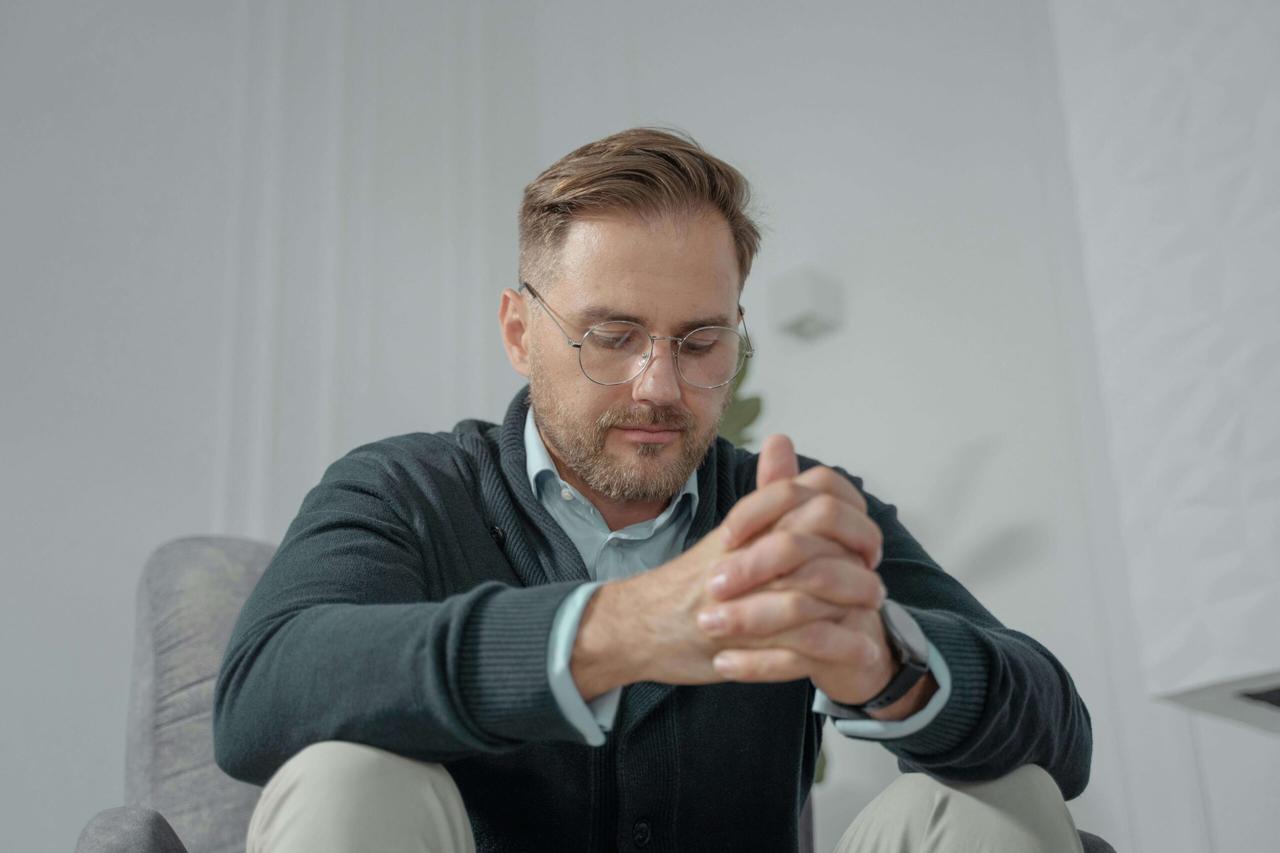 Chronic Anxiety, image of a sad man sitting on a couch, looking down, with his arms resting on his knees and his hands together.