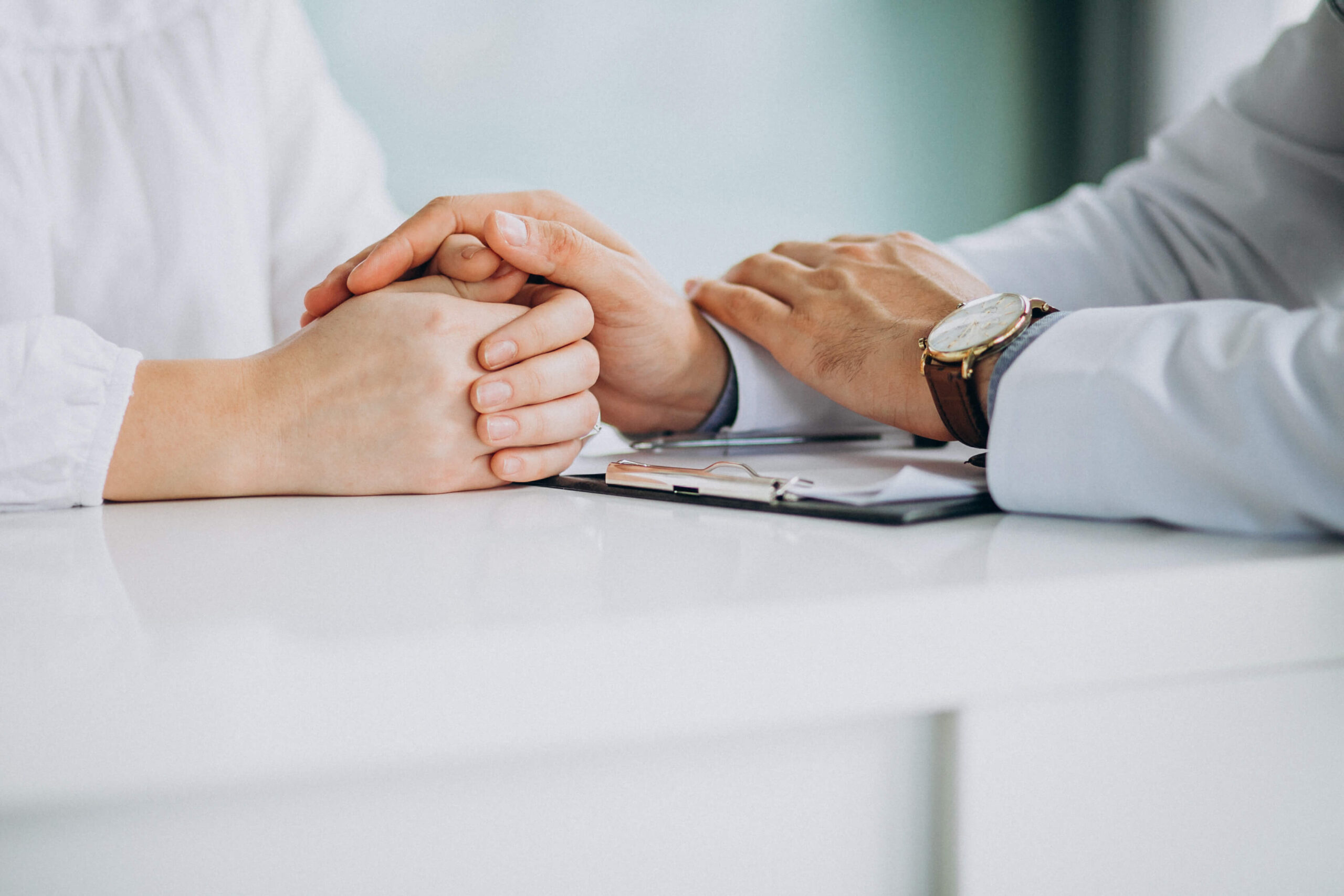 Anxiety, image of a doctor’s hands resting on a female patient’s hands on a white desk surface.