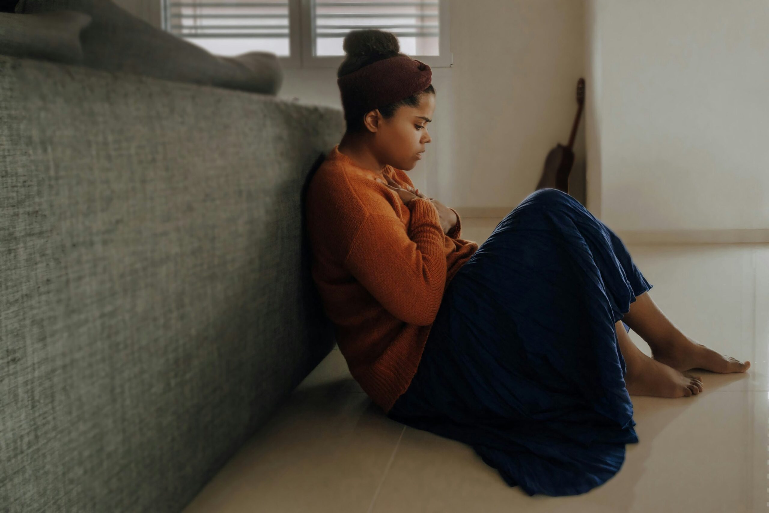 Treating depression, image of a young woman wearing an orange sweater and long blue skirt sits on the floor behind a gray couch with a sad expression on her face.
