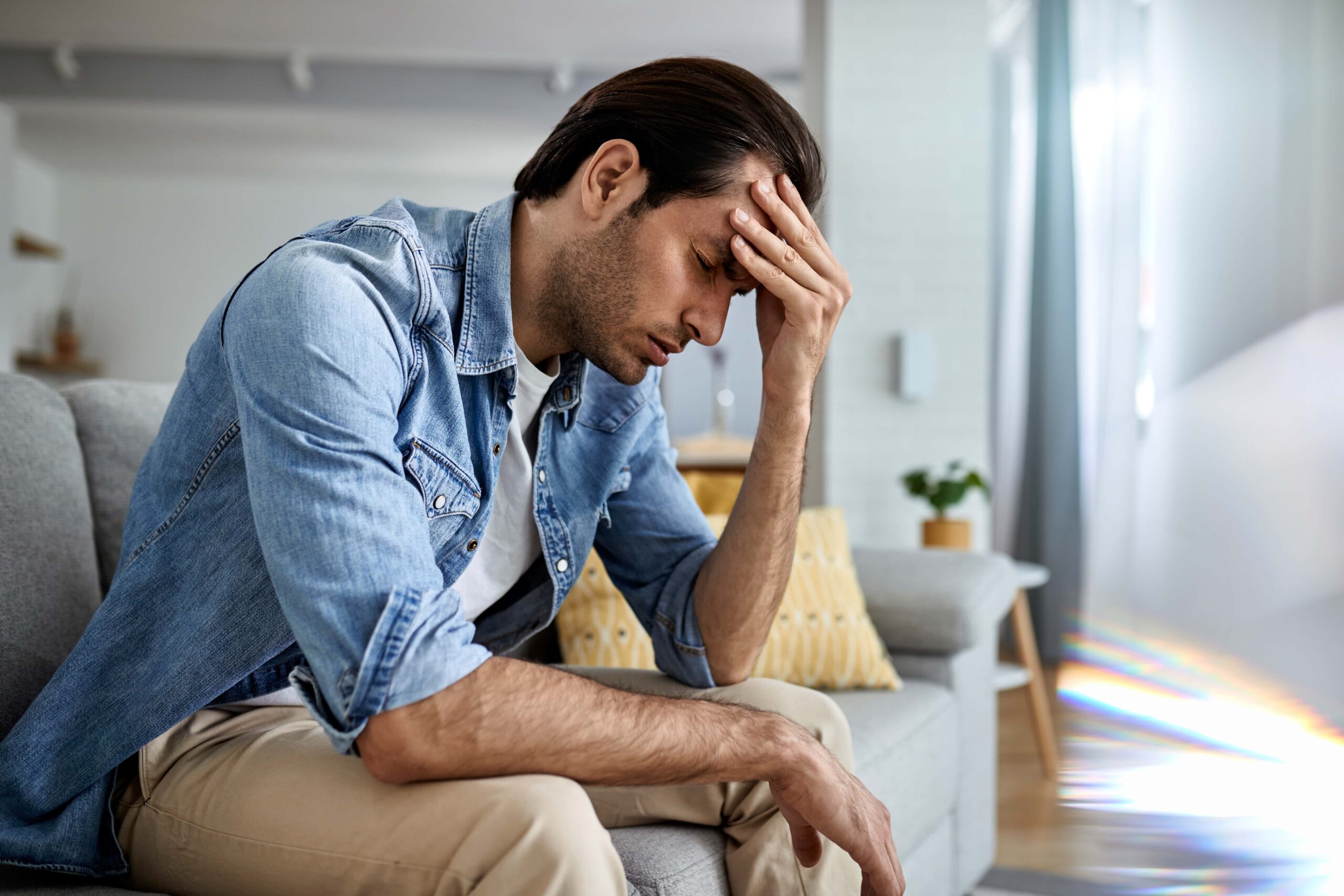 Treating depression, image of a man wearing a jean button-down shirt and khaki pants, sits on a gray couch with his left hand on his forehead and his right arm resting on his right leg with a painful expression on his face.