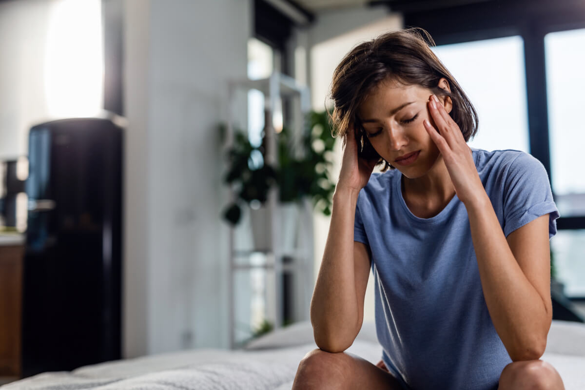 Adrenal Stress, image of a woman with dark, short hair, wearing a light blue t-shirt, sitting on the edge of a bed with her hands pressed against the sides of her face with an expression of pain.