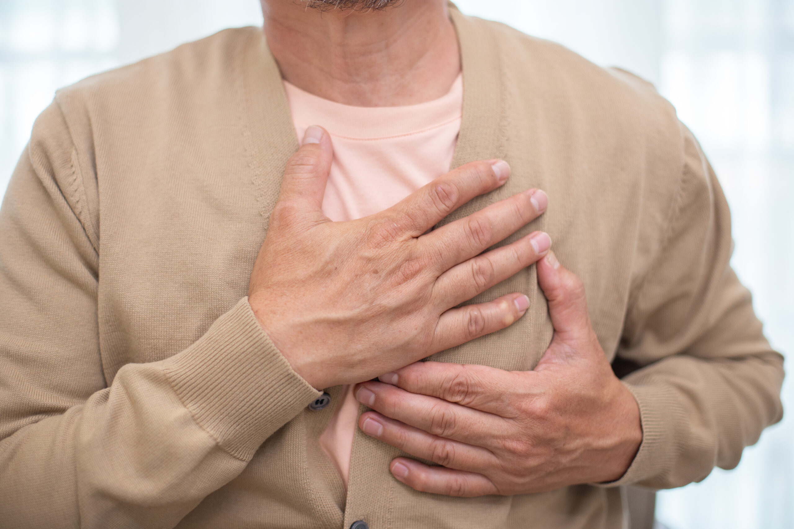 High Blood Pressure (Hypertension), image of a man, wearing a tan cardigan sweater and a pink undershirt, grasps his chest with both of his hands.