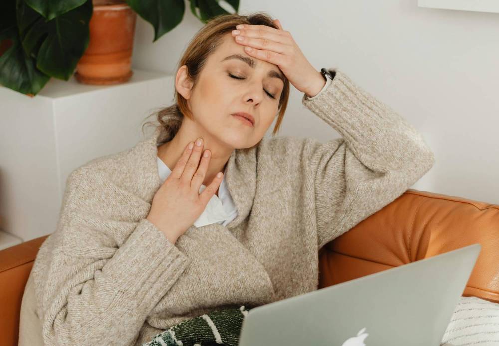 Chronic Inflammatory Response Syndrome (CIRS), image of a woman sitting on a couch wearing a tan sweater with one hand on her neck and another hand on her forehead and a laptop in her lap